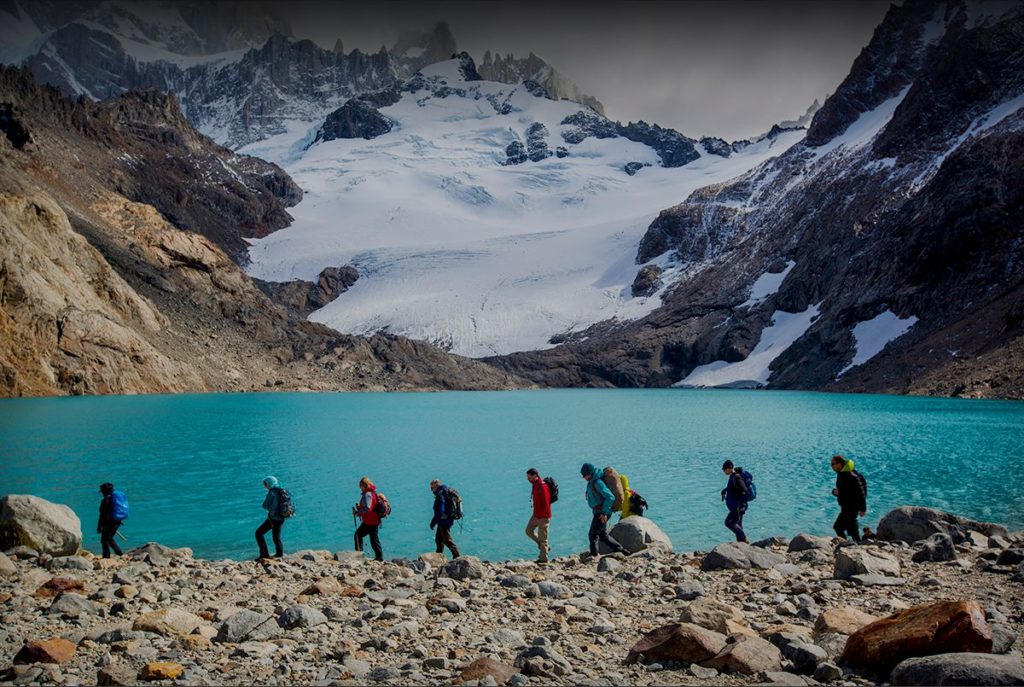 a group of people walking on a rocky shore with a body of water and snow covered mountains