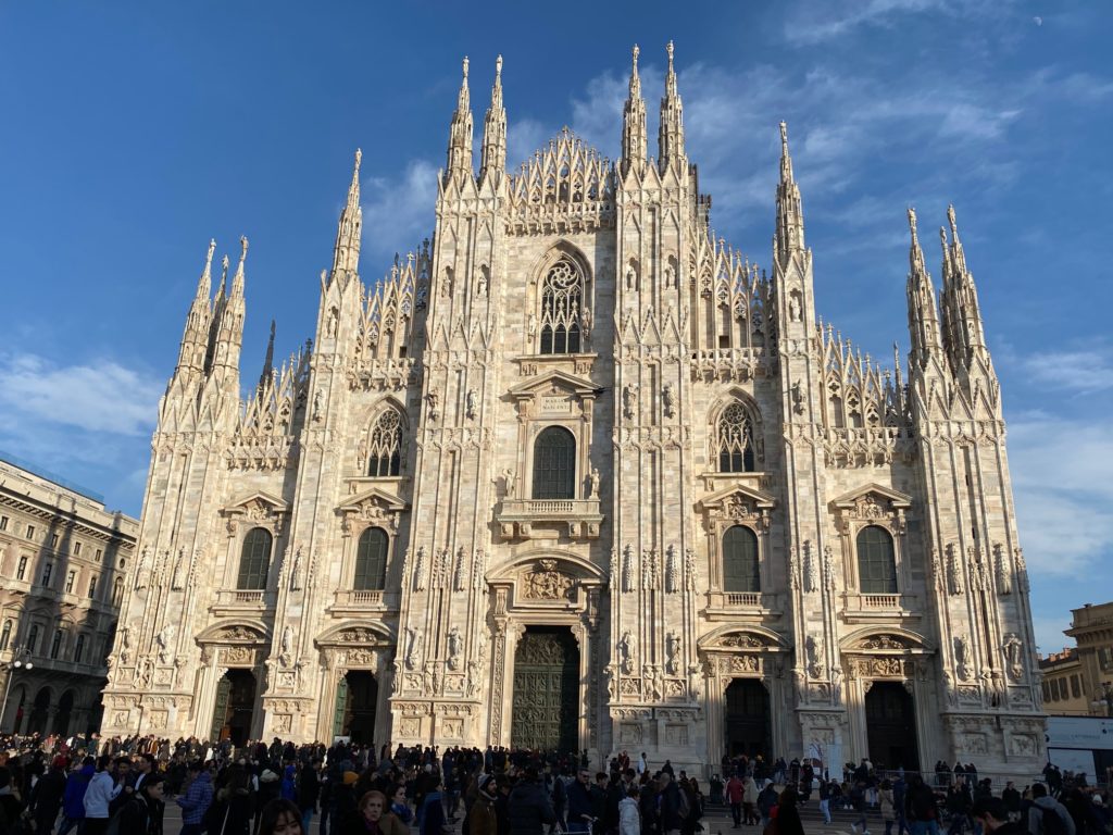 a large white building with many people in front with Milan Cathedral in the background