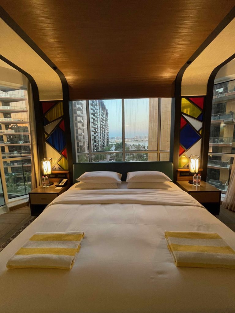 a bed with white sheets and yellow towels in a room with large windows